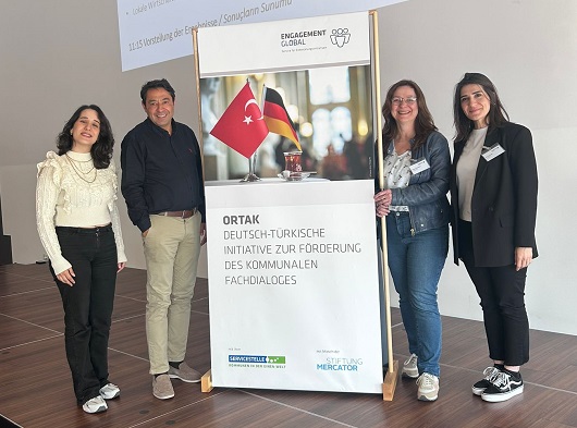 Eine Gruppe von vier Personen steht neben dem Banner von Ortak/Engagement global und lächelt freundlich in die Kamera
