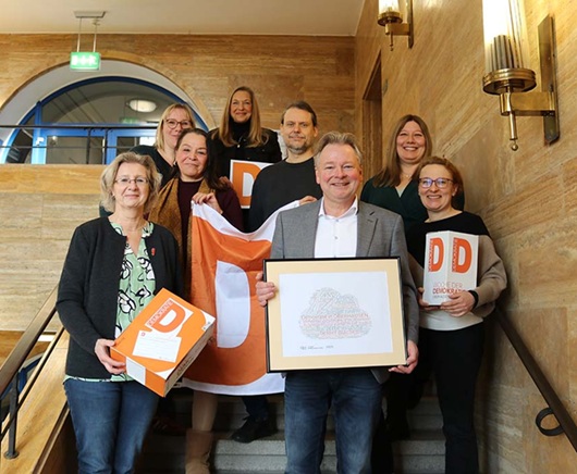 Eine Gruppe von Menschen steht auf einer Treppe und lächelt in die Kameras. Sie halten das orange D für Demokratie hoch. (Foto: Stadt Oberhausen/Tom Thöne)