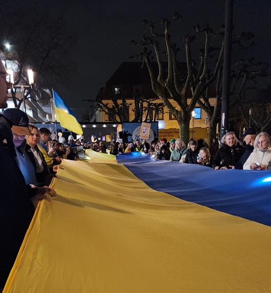 Menschen tragen gemeinsam eine riesige Ukraine-Flagge vor einem schwarzen Himmel