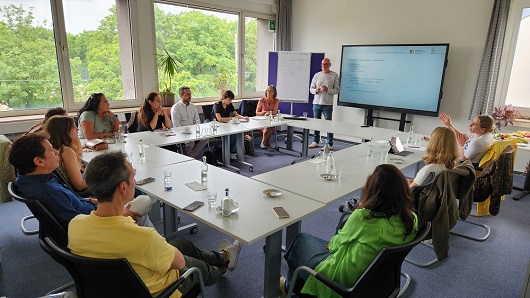 Menschen sitzen um einen U-förmigen Tisch in einer Arbeitssituation, vorn steht ein Mann und hält eine Präsentation