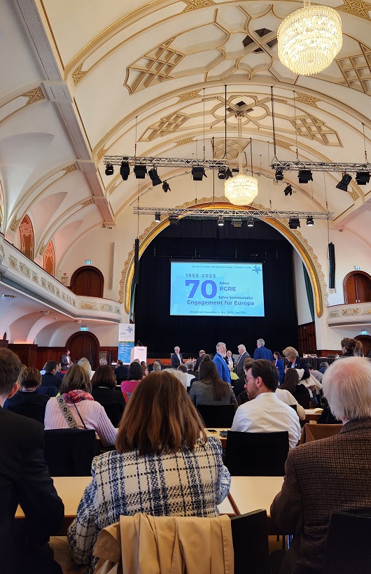 Bild von der Delegiertenversammlung, Anwesende von hinten, vorn die Präsentation mit dem Titel „70 Jahre RGRE – 70 Jahre kommunales Engagement für Europa“