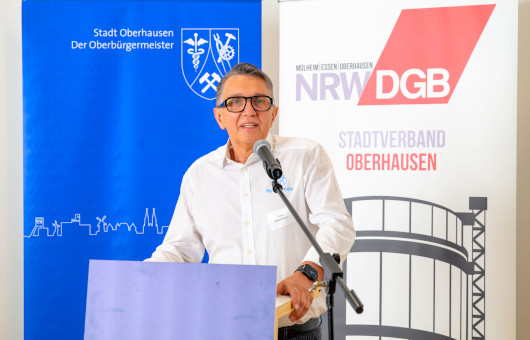 Mann im weißen Hemd mit Brille spricht in ein Mikrofon. Im HIntergrund links ein blaues Roll-Up und rechts ein weißses Roll-Up mit dem Gasometer Oberhausen